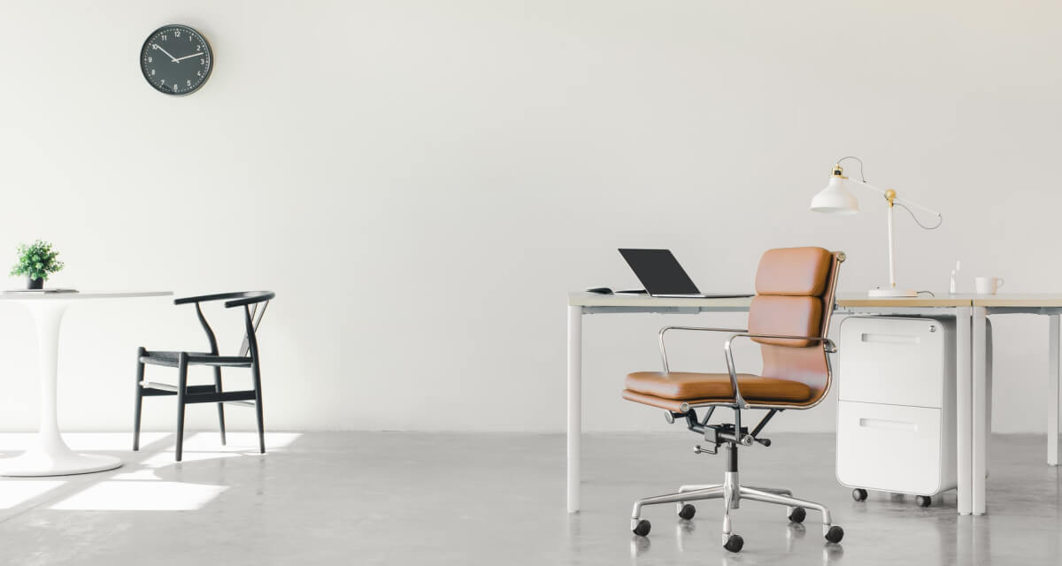 Eine minimalistische Büroeinrichtung von Möbelhaus besteht aus einem weißen Schreibtisch, einem braunen Drehstuhl, einem Laptop, einer weißen Schreibtischlampe und einem kleinen Aktenschrank. Links steht ein runder Tisch mit zwei Stühlen, an der weißen Wand darüber hängt eine schwarze Uhr. Eine kleine Pflanze schmückt den Tisch. Weitere Informationen finden Sie in unserem Sigmaringer Küchenstudio.