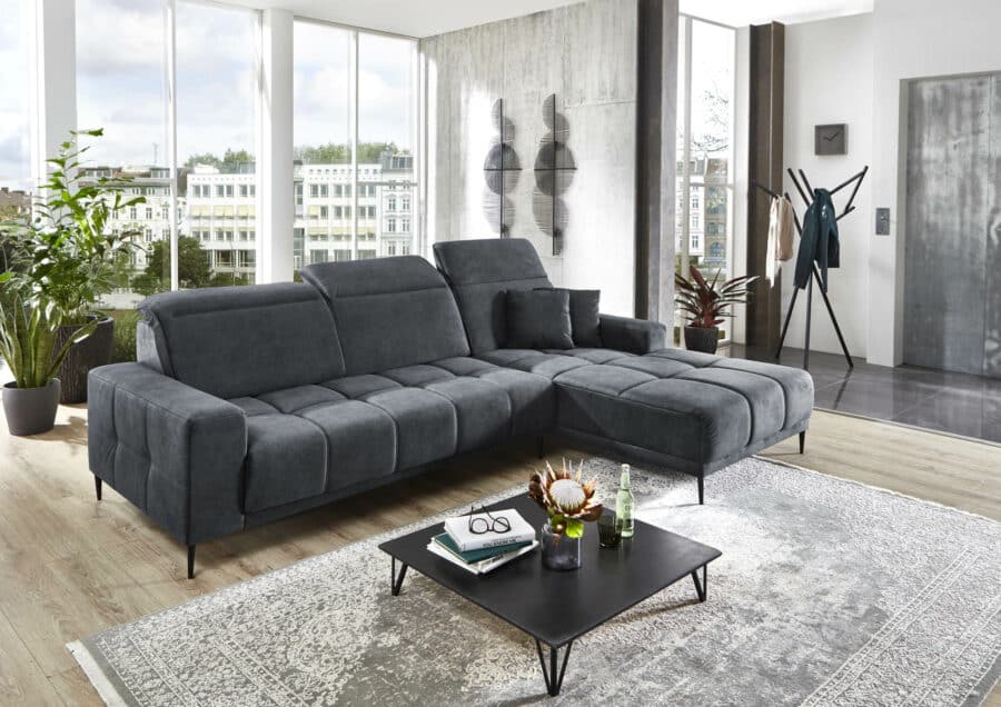 Ein modernes Wohnzimmer mit einer Polstergruppe 8795 Livorno Type 001, einem schwarzen Couchtisch mit Dekoartikeln und einem strukturierten grauen Teppich. Große Fenster lassen Tageslicht herein, während Pflanzen und Wanddekoration für Charme sorgen. Angrenzend befindet sich ein offener, zeitgenössischer Raum, der an das elegante Design eines nahegelegenen Küchenstudios erinnert.