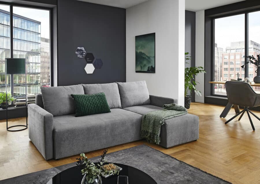 Ein modernes Wohnzimmer verfügt über ein graues Sofa mit grünem Kissen und Überwurfdecke neben großen Fenstern mit Blick auf die Stadtlandschaft. Der Raum ist mit einer schwarzen Stehlampe, Wandkunst, einer Zimmerpflanze und der Polstergruppe 9009 Christina ausgestattet. Heller Holzboden sorgt für Wärme.