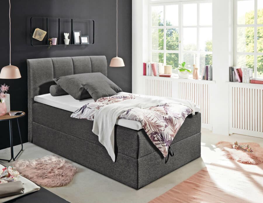 Ein gemütliches Schlafzimmer mit einem Einzelbett, dem 8018 Granada Boxspringbett mit grau gepolstertem Kopfteil und Untergestell von Möbel Sigmaringen. Das Bett ist hübsch gemacht mit weißen Laken und einer Bettdecke mit einem rosa-grauen Blättermuster. Das Zimmer hat eine schwarze Akzentwand, große Fenster, einen Beistelltisch und hängende Pendelleuchten.