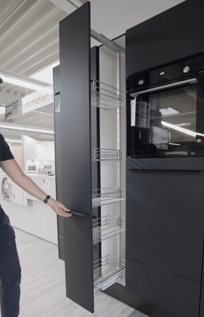 Eine Person zieht aus einem modernen Küchenschrank eine vertikale Vorratsschrankeinheit mit Drahtregalen heraus. Das elegante Design weist eine mattschwarze Oberfläche auf, die den integrierten schwarzen Ofen rechts ergänzt. Dieses Küchenstudio in Sigmaringen besticht durch eine minimalistische Ästhetik mit hellem Holzboden, perfekt für jedes moderne Zuhause.