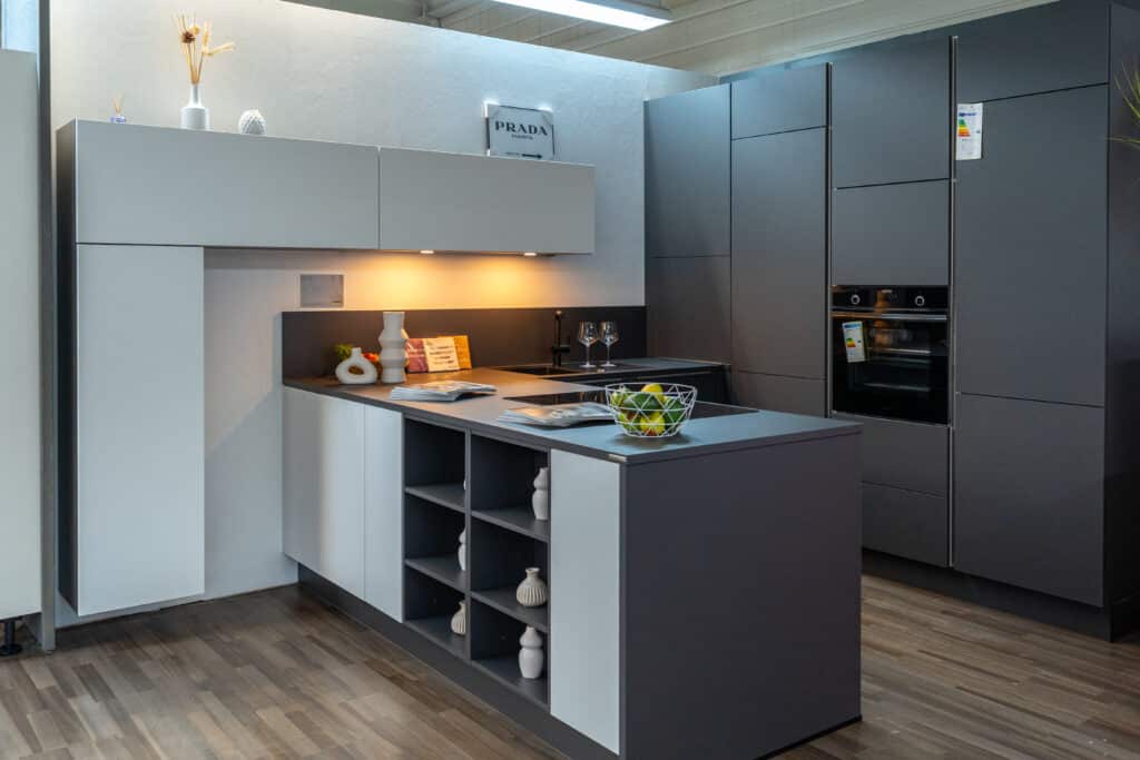 Eine moderne Küche mit elegantem Design und grau-weißen Schränken, entworfen von einem renommierten Küchenstudio in Sigmaringen. Die Kücheninsel verfügt über eingebaute Regale für Vasen und Dekoartikel. Edelstahlofen und andere Geräte sind in die hohen Schränke integriert. Warme Beleuchtung unter den Schränken rundet den Look ab.
