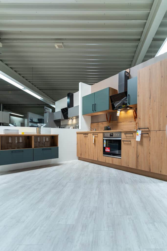 Ein modernes Küchenstudio in Sigmaringen verfügt über elegante Schränke in Blaugrün und Holzoptik, weiße Arbeitsplatten und Geräte aus Edelstahl, darunter ein Backofen und eine Mikrowelle. Der Raum besticht durch ein zeitgenössisches, minimalistisches Design mit einer hellen, sauberen Atmosphäre.