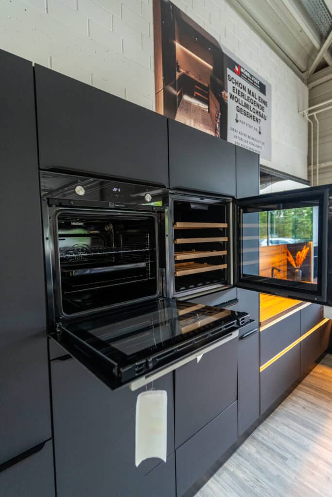 Ein modernes Küchenstudio mit eleganten, dunklen Schränken. In die Schränke sind ein offener Ofen und eine angrenzende offene Schublade eingebaut. Darüber hängt ein großes, farbenfrohes Foto, während natürliches Licht durch ein Fenster auf der rechten Seite hereinströmt. Der Boden hat eine helle Holzmaserung und schafft ein stilvolles Ambiente, wie man es in jedem Möbelhaus finden würde.