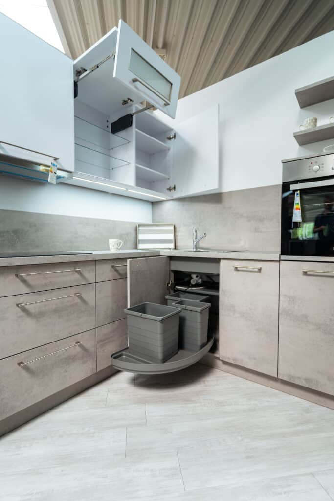 Moderne Küche mit grauen Schränken und Schubladen, offenen Oberschränken und ausziehbarem Eckschrank mit zwei grauen Behältern. Schlichte graue Arbeitsplatten, eingebauter Backofen und eine Keramikspüle mit einem Griff. Heller Holzboden und eine Holzdecke darüber. Entworfen vom renommierten Küchenstudio in Sigmaringen.