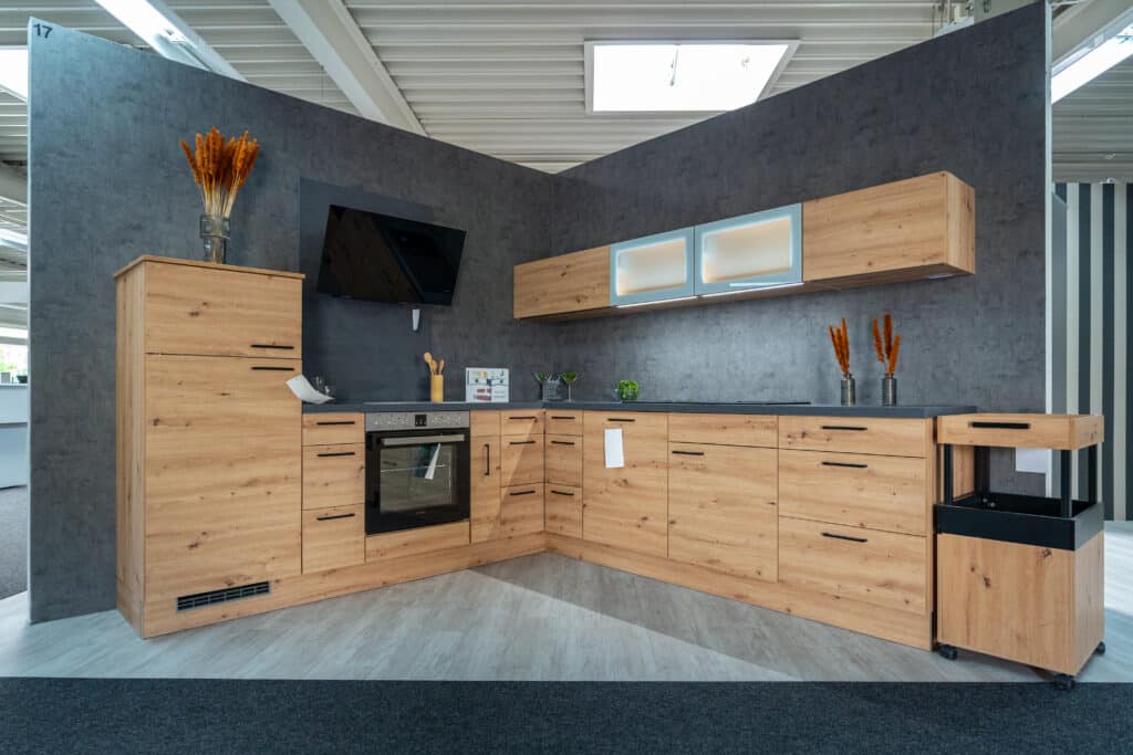Eine moderne Eckküche verfügt über helle Holzschränke mit dunkler Arbeitsplatte. Zu den Geräten gehören ein eingebauter Ofen und ein Herd. Über dem Herd befindet sich ein schwarzer, an der Wand montierter Fernseher. Zu den dekorativen Elementen, die von einem örtlichen Küchenstudio geliefert wurden, gehören Töpfe mit Trockenblumen.