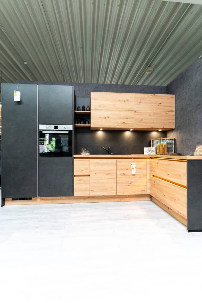 Eine moderne Küche mit hellen Holzschränken und schwarzen Akzenten, mit eingebautem Ofen, schwarzem Kühlschrank und Unterschrankbeleuchtung. Dieses Küchenstudio verfügt über eine Wellblechdecke und einen hellgrauen Boden, was eine elegante und doch industrielle Atmosphäre schafft.