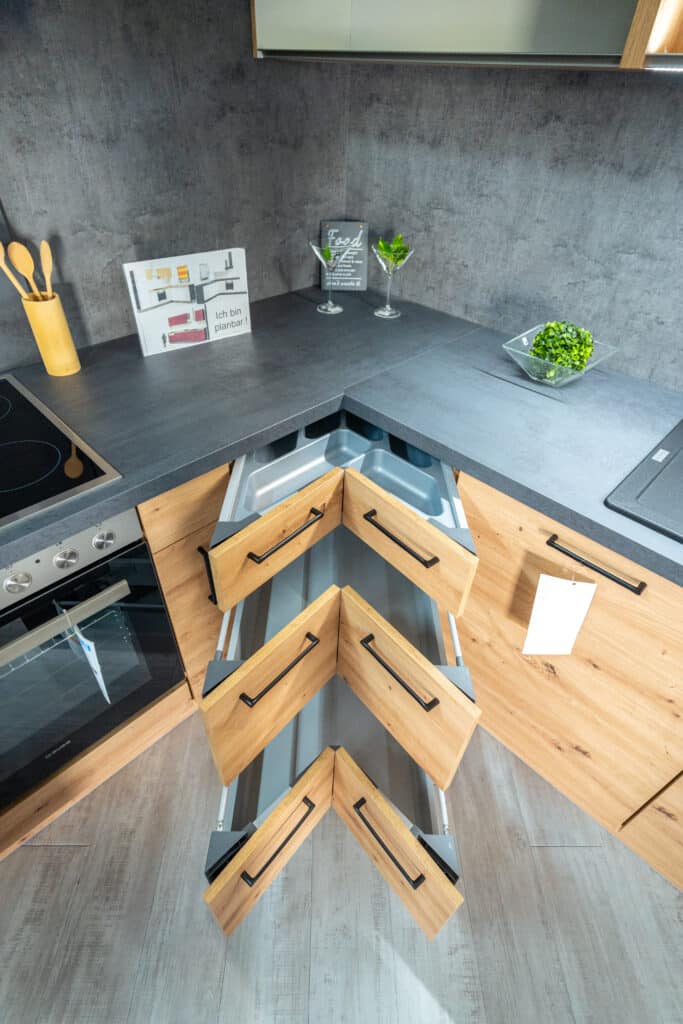 Eine moderne Küche in Sigmaringen mit Holzschränken und dunkler Arbeitsplatte von einem renommierten Möbelhaus. Der Eckschrank verfügt über ein einzigartiges Design mit vier ausziehbaren Schubladen. Auf der Arbeitsplatte stehen zwei Martinigläser, ein dekoratives Schild und eine kleine Pflanze. Auf der anderen Seite befinden sich ein Kochbuchständer und ein Messerblock.