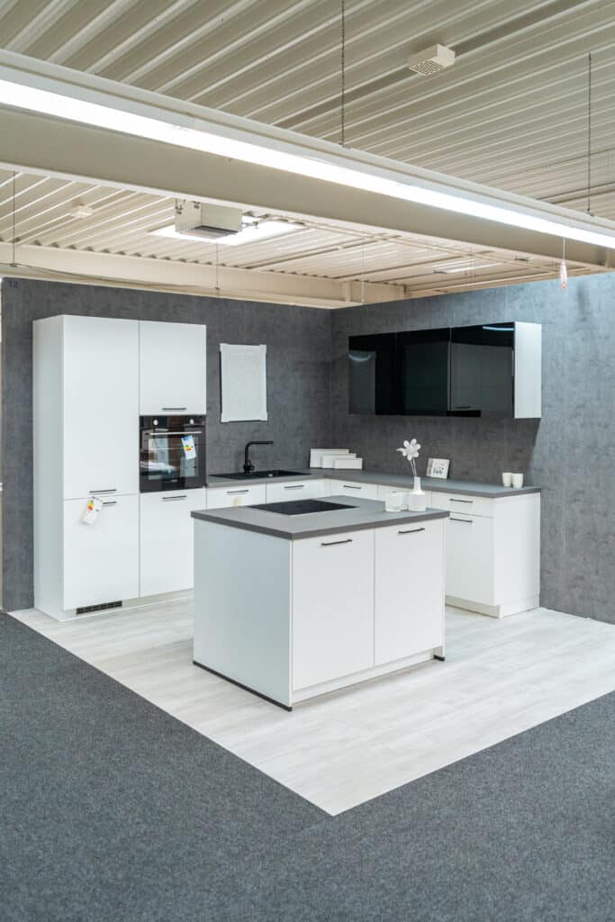 Ein moderner Küchenausstellungsraum im Möbelhaus Sigmaringen mit eleganten weißen Schränken, einer zentralen Kücheninsel mit schwarzer Arbeitsplatte, eingebautem Ofen und minimalistischen schwarzen Wandschränken. Die grauen Wände mit Betonstruktur bilden einen kontrastreichen und dennoch stilvollen Hintergrund.