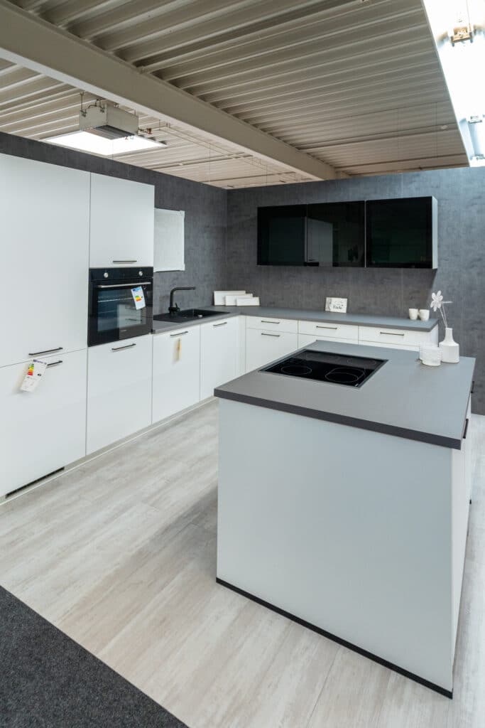 Modernes Kücheninterieur von Küchenstudio in Sigmaringen mit weißen Schränken, einer dunklen Arbeitsplatte auf der Kücheninsel und einem eingebauten schwarzen Ofen. Der Boden ist mit hellem Holz ausgelegt, während Deckenbeleuchtung das elegante Design erhellt. Die Kücheninsel verfügt über ein Kochfeld und minimalistisches Dekor, was für einen klaren Look sorgt.