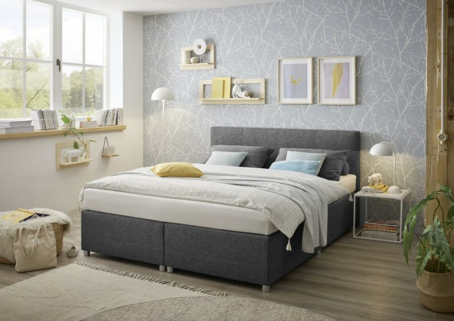 Ein modernes Schlafzimmer mit einem großen 9066 Luanda Boxspringbett mit weißer und hellblauer Bettwäsche. Das Zimmer hat hellgraue Tapeten mit Naturmotiven, Holzböden und ein großes Fenster. Wandregale halten dekorative Gegenstände und eine Topfpflanze auf dem Boden verleiht einen Hauch von Sigmaringen-Charme.