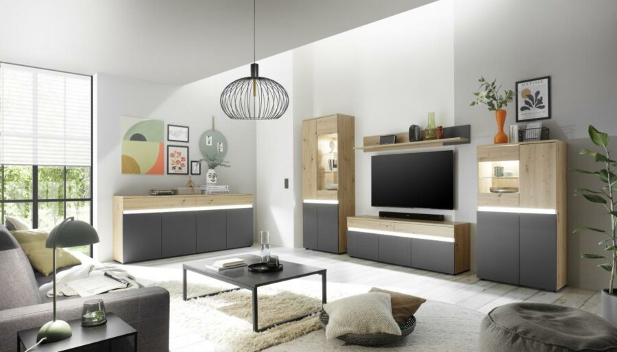 Modernes Wohnzimmer mit minimalistischem Dekor. Zur Ausstattung gehören ein großer Flachbildfernseher an einer 9069 Lille Wohnwand, Wandregale mit Dekoartikeln, ein graues Sofa, ein schwarzer Couchtisch und ein flauschiger weißer Teppich. Natürliches Licht strömt durch große Fenster.