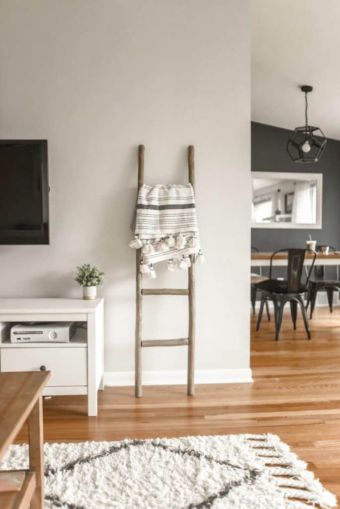 In einem minimalistischen Wohnzimmer in Sigmaringen lehnt eine Leiter mit einer darüber gehängten gestreiften Decke an einer weißen Wand. Daneben ist eine weiße TV-Konsole von Möbel mit einer Topfpflanze und elektronischen Geräten zu sehen. Im Hintergrund ist ein Essbereich mit einem schwarzen Tisch und Stühlen zu sehen.