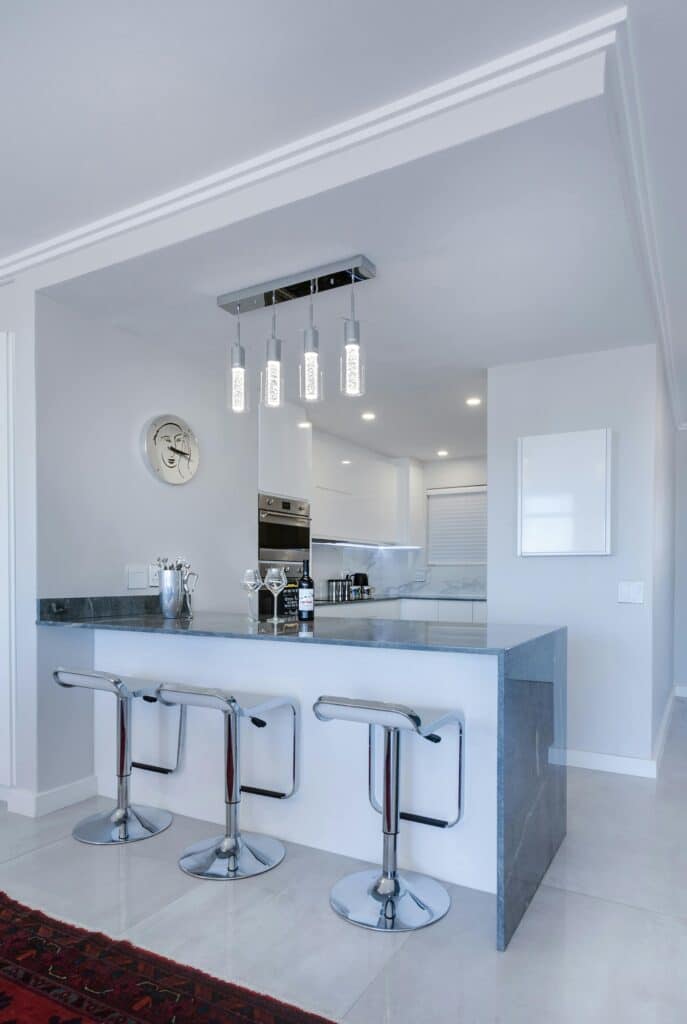Moderne Küche mit minimalistischem Design. Mit einer Farbpalette aus Weiß und Grau, Hängelampen und Geräten aus Edelstahl. Im Vordergrund steht eine graue Kücheninsel mit drei eleganten Metallhockern. Der Boden ist hell, mit einer teilweise sichtbaren roten Teppichkante aus der Küchenstudio Möbel-Kollektion.