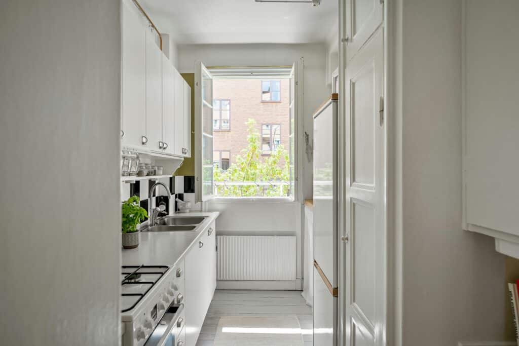 Eine schmale Küche mit weißen Schränken, einer Edelstahlspüle, einem Gasherd und einem Backofen. Ein kleines Fenster über einem Heizkörper lässt den Raum mit natürlichem Licht durchfluten und gibt den Blick auf ein Backsteingebäude und Grünflächen draußen frei. Eine grüne Pflanze steht auf der Theke und verleiht diesem gemütlichen kulinarischen Möbelensemble Leben.